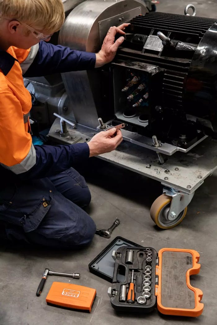 A mechanic in safety gear repairs industrial machinery using a toolset indoors.